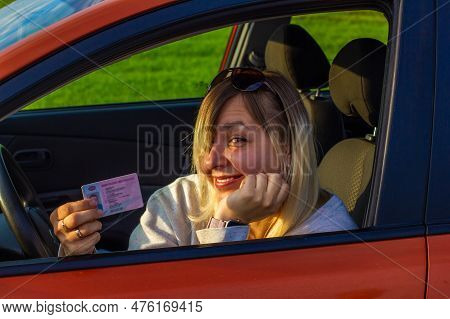 A Happy Woman Sits Behind The Wheel Of A Car And Shows Her New Drivers License. A Woman Is Smiling C
