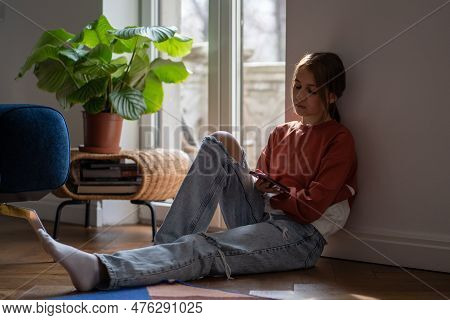 Cyberbullying, Negative In Social Media. Upset Teen Girl Sitting On Floor Near Window Using Smartpho