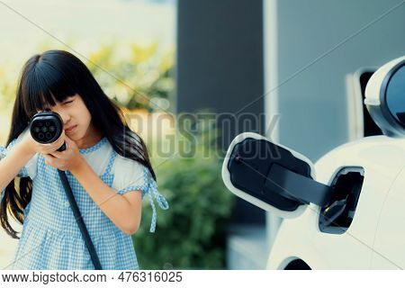 A Playful Girl Holding And Pointing An Ev Plug, A Home Charging Station Providing A Sustainable Powe