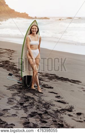 Healthy Smiling Fit Woman In Bikini Stands With Surfboard, On Black Beach By The Ocean. Extrime Wate