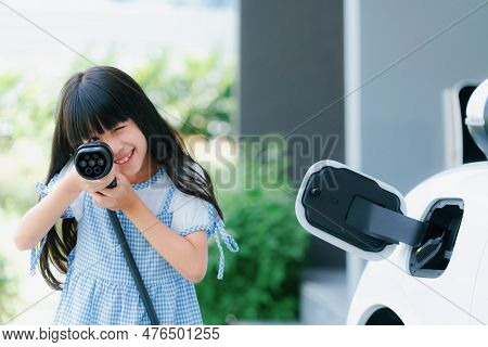 A Playful Girl Holding And Pointing An Ev Plug, A Home Charging Station Providing A Sustainable Powe