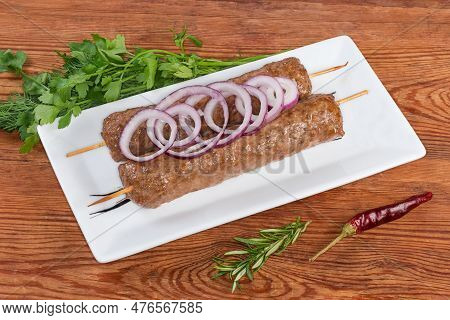Two Baked Lula Kebab Skewered On Wooden Skewers Decorated With Red Onion Rings On The White Rectangu
