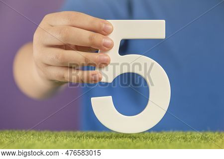 Number 5 In Hand. A Hand Holds A White Number Five On Green Grass And A Purple Background, Five Perc