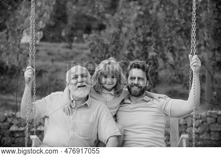 Grandfather With Son And Grandson Swinging Together In Summer Garden On Swing Outdoors. Active Leisu