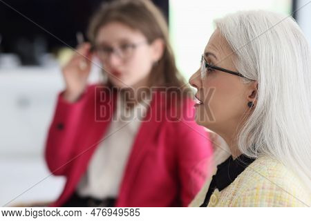 Serious Female Employees Discuss Working Details Sitting On Chairs In Light Office. Young And Elderl