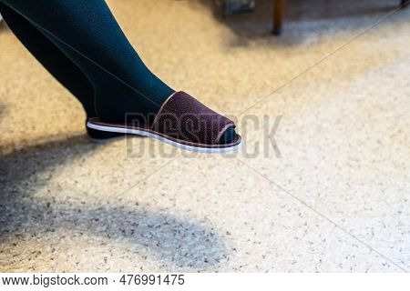 Woman's Leg In Dark Tights And House Slipper In Apartment Lit By Electric Light Over Linoleum Floor