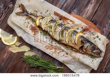 Whole baked mackerel or scomber fish with lemon and greens on paper dark wooden background top view