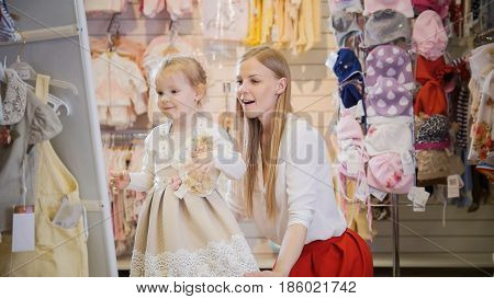 Shopping for kids - blonde little girl with mommy chooses dress in store of kids clothes, telephoto