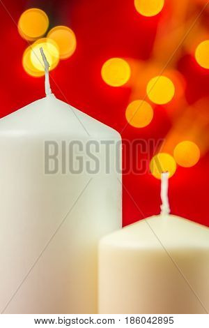 Two white christmas wax candles on red background with bright yellow lights playing with lights burning candles