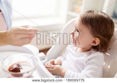 family, food, eating and people concept - mother with puree and spoon feeding little baby sitting in highchair at home