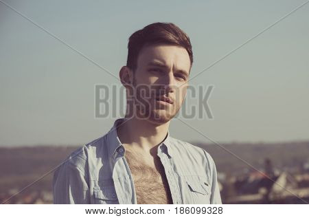 Sexy young caucasian handsome man or model with unshaven face and stylish hair haircut posing in unbutton shirt with hairy naked chest on sunny summer day on grey sky background. Male beauty
