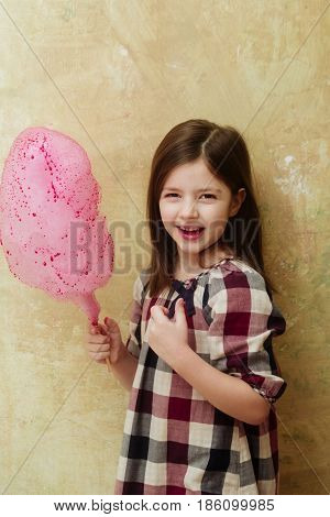 Happy adorable girl small little child in plaid dress smiling with delicious pink cotton candy sweet sugar spun candyfloss on stick on beige background. Unhealthy food or snack
