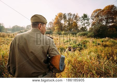 Dyatlovichi, Belarus - October 2, 2016: Reenactor Dressed As Russian Soviet Red Army Soldier Of World War II Hidden Sitting And Aiming Rifle Weapon In Grass In Autumn Forest