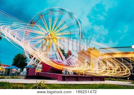 Motion Blurred Effect Around Of High Speed Rotating Illuminated Attraction Feature In City Amusement Park. Ferris Wheel On Summer Evening Blue Sky Background.