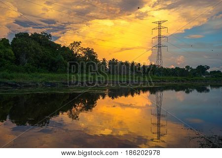 High voltage electric transmission tower with reflection in the river during golden hour sunset.