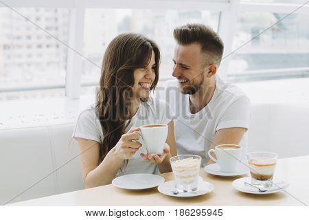 Smiling young couple on a date in a cafe. They are sitting on a big white sofa near panoramic windows and drinking coffee or chocolate
