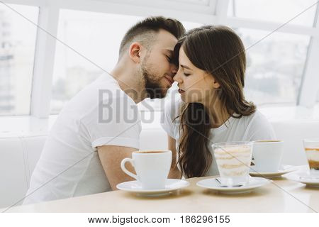 Attractive happy young couple on a date in a cafe having some tasty coffee or chocolate. Handsome guy is trying to kiss his girlfriend