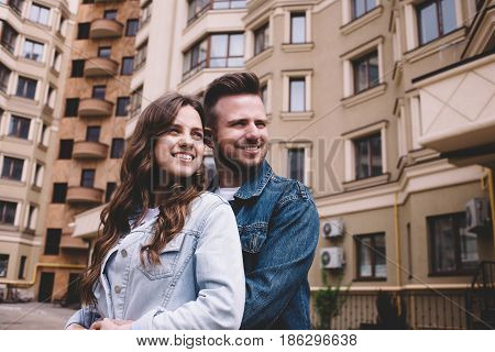 Positive and awesome young couple on a date. They are standing among huge nice buildings in a warm hugs and looking somewhere. Close up