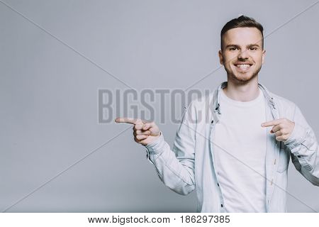 Happy smiling man standing on white background. Pleasant guy pointing aside with forefinger at something really great. Place for advert. Close up. Isolated