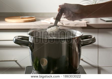 Female hand holding spoon with rice above metal pan on hotplate in kitchen