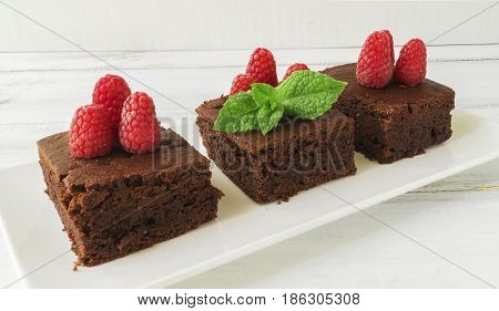 Three pieces black bean brownie cake decorated with raspberry and mint on white rectangular plate on white wooden table selective focus horizontal
