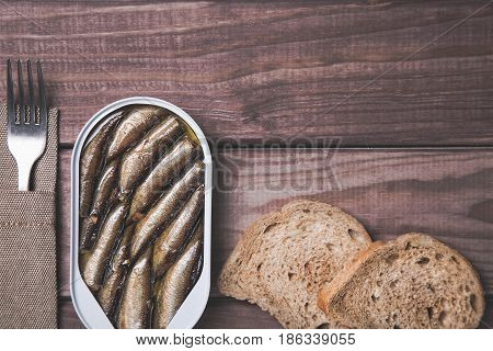 Tin can of sprats, sardines with bread pieces and fork on wooden table. Top view and free space.