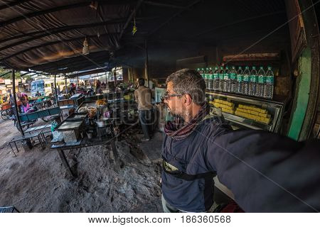 Tourist taking selfie at street food stall in India bus station while traveling. Fish eye view with indian sweets in foreground.