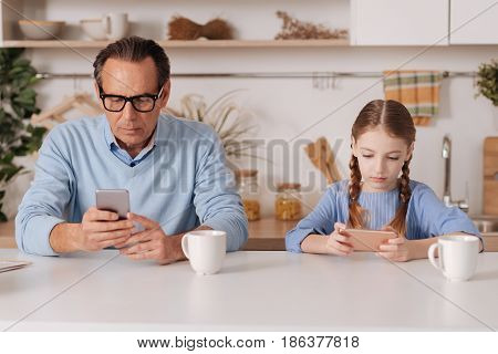 Forever online disease. Retired addicted aging pensioner sitting indoors and using smartphone with granddaughter while surfing the Internet and checking social media profiles
