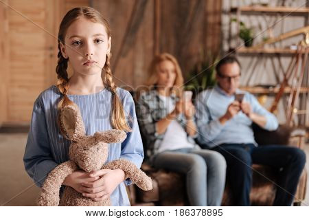 Feeling neglected. Neglected puzzled little girl waiting for parents attention at home and holding toy while adults using devices and paying no attention