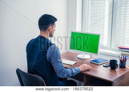 Young man office worker sitting at computer with Chromakey and looking on monitor. Office concept with working environment. Back view. Copyspace on screen