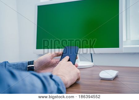 Man in office typing on telephone. computer with Chromakey is on background. Office concept with working environment. Copyspace on screen