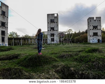 A young woman with long blond hair stands against the background of three half-ruined towers on a cloudy day. The girl raised her face to the sky and the wind blows her hair