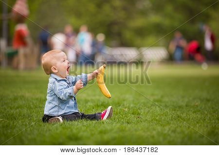 Adorable toddler boy with banana sitting on the green grass in the city park and shouting. Child walking in summer park.