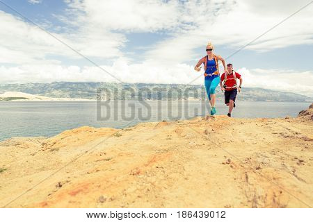 Couple runners running with backpacks on rocky trail at seaside and mountains. Young woman and man trail running on mountain path looking at beautiful inspirational landscape view.
