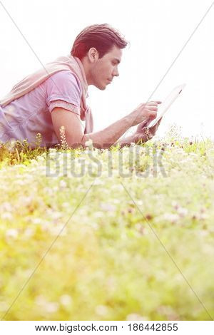 Side view of young man using digital tablet while lying on grass against clear sky