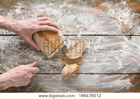 Baking background. Hands of baker cutting bread loaf with knife on rustic wooden table sprinkled with flour.