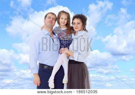 Happy young family, mom dad and little daughter.Parents keep the girl in her arms , and she hugs their neck.On the background of summer blue sky and fluffy clouds.