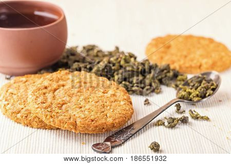 Dry leaves of green tea in a glass jar. Nearby tea leaves in a teaspoon and ceramic tea-things. Composition on a wattled napkin. The cover banks is open several tea leaves are scattered.