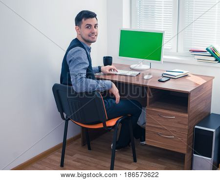 Happy office worker smiling and looking at the camera. He sits at computer with Chromakey. Office concept with working environment. Copyspace on screen