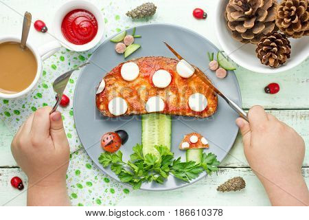 Child eat funny brunch from toast with tomato sauce cheese and cucumber shaped mushroom on vegetables greens forest glade. Fun with food composition top view