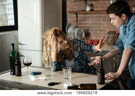 Feel depressed. Angry teenager standing in semi position opposite his mother leaning on table while gesticulating