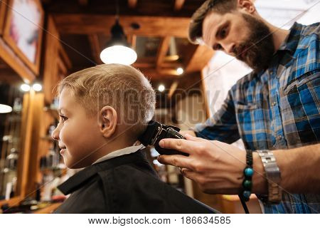 Cutting hair. Nice bearded male hairdresser holding a hair cutting machine and cutting his clients hair while standing behind him