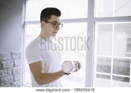 Young concentrated sportsman doing workout up and down with left arm with dumbbell while standing near a big and white window. Profile view. Close up