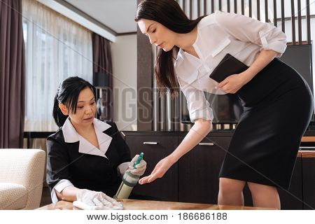 It is not clean. Professional nice housekeeping manager holding the tablet and leaning over the table while checking the work of the hotel staff