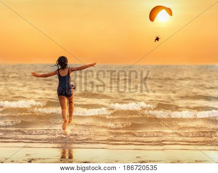 The girl runs into the sea pretending to parody the paramotor flying in the evening the sun is falling the concept of relaxation.