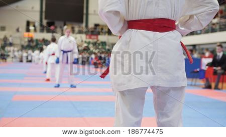 The athlete stands in a white kimono with a red belt preparing for a fight with an opponent in a white kimono with a blue sash and blue fighting gloves. For springa watching the judge in a black suit with a red flag. Competitions in karate among young ath