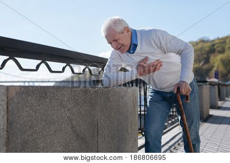 Without support. Mirthless calm scared man holding his heart standing by stick and expressing ache on his face