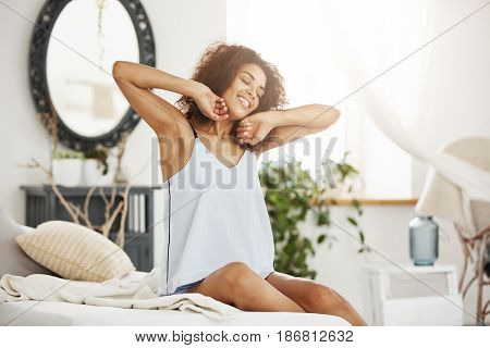 Young happy beautiful african girl in sleepwear stretching smiling with closed eyes sitting on bed at home.