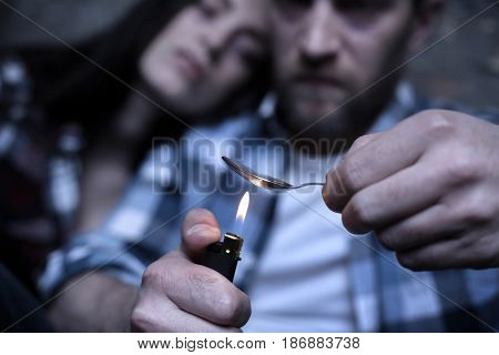 Severe physical and mental suffering. Skilled concentrated young junkies siting in the darkness while heating drug dose and using spoon and lighter