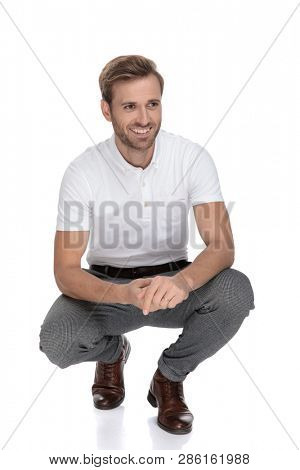 crouched casual man smiles and holds palms together while looks to side on white background